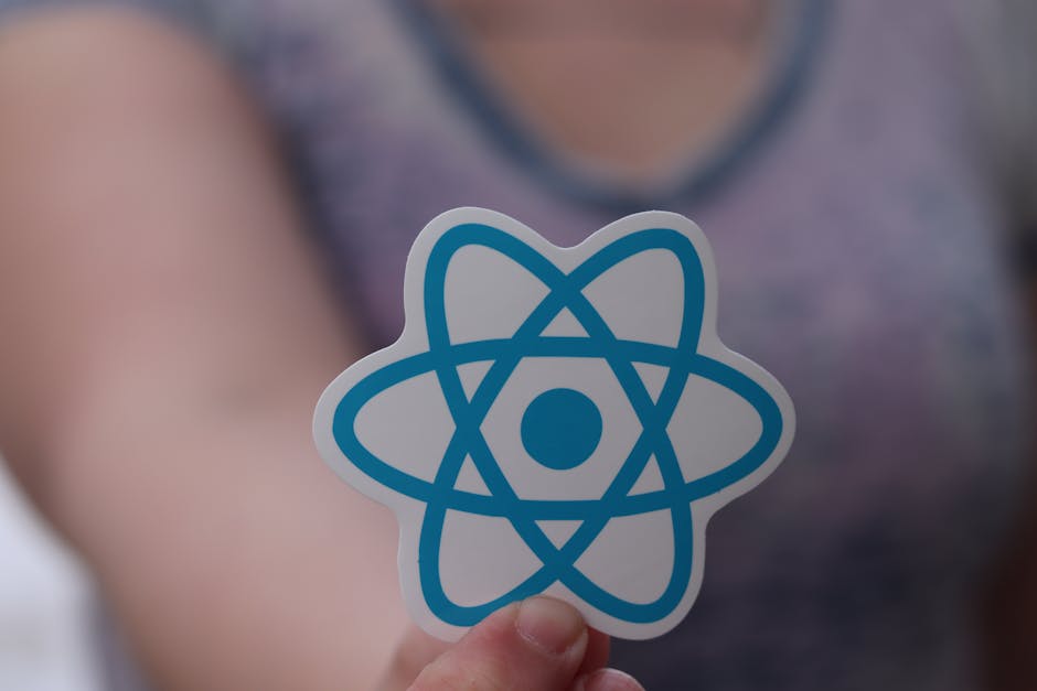 Close-up of a person holding a white and blue sticker featuring an atomic model symbol, with a blurred background of a person wearing a purple sleeveless top and a grey shirt. The focus is on the stic